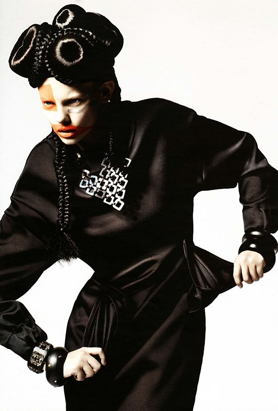 Woman in avant-garde black satin dress, elaborate braided headpiece, and statement necklace poses in a fashion forward studio portrait.