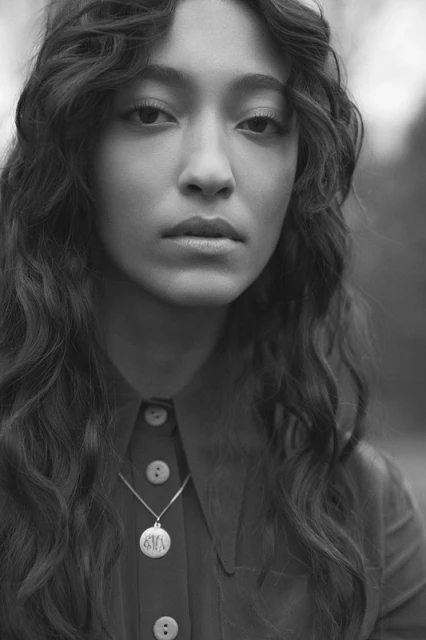 Monochrome portrait of a young woman with wavy long hair, wearing a collared shirt and pendant necklace against a blurred background.