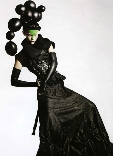 Fashion photo featuring a single model with green face makeup and black balloons, wearing a black gown and gloves against a white backdrop.