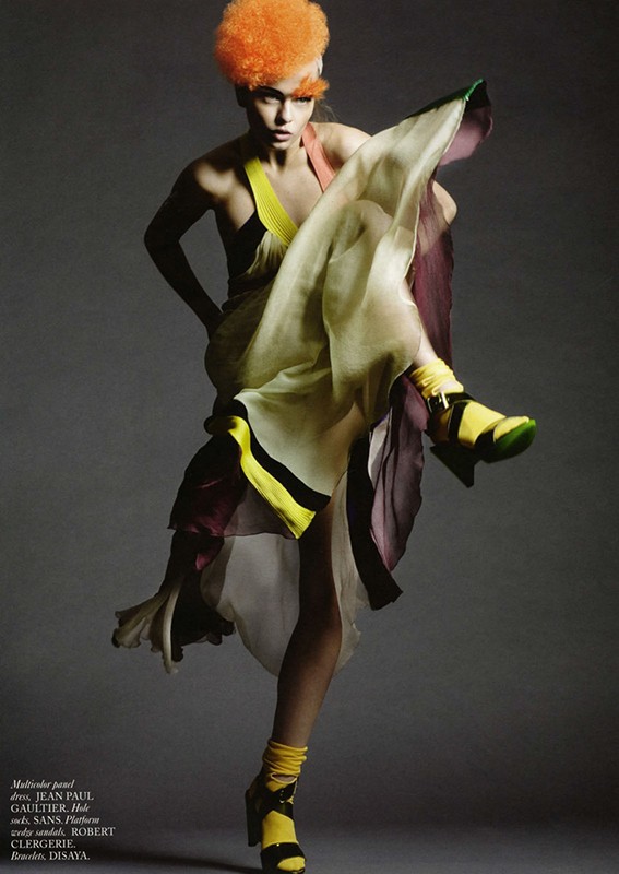 Fashion editorial shot of a woman with orange hair in a flowing multicolor dress and yellow platform sandals, posing in a photo studio.