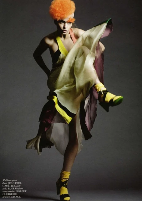 Fashion editorial shot of a woman with orange hair in a flowing multicolor dress and yellow platform sandals, posing in a photo studio.