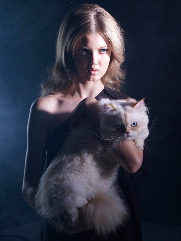 Portrait of a blonde woman in a black dress holding a fluffy white cat against a dark background with soft, diffused lighting.