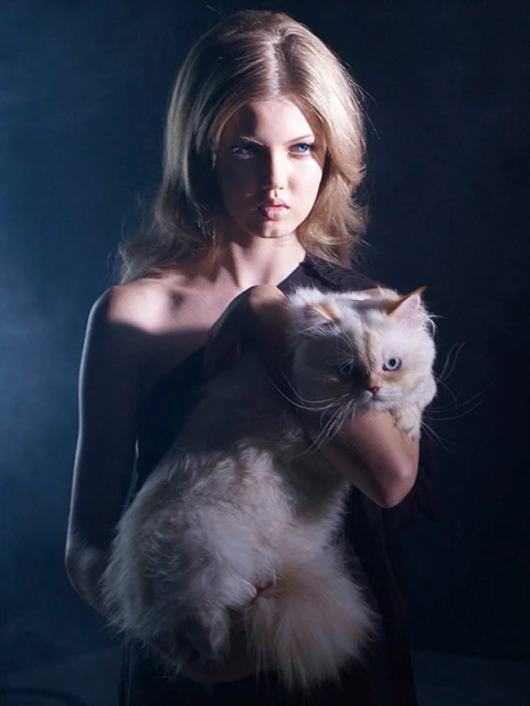 Portrait of a blonde woman in a black dress holding a fluffy white cat against a dark background with soft, diffused lighting.