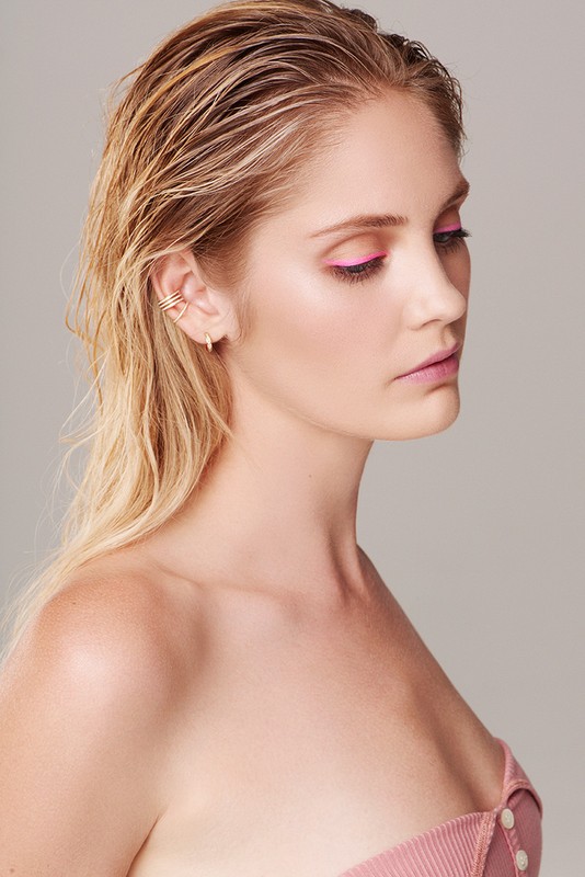 Close-up profile of a blonde woman with pink eye makeup, gold earrings, and a mauve strapless top against a neutral background.