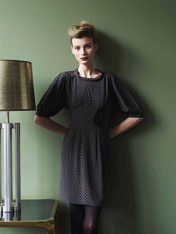 A woman in a vintage-inspired dress poses indoors with soft lighting next to a modern lamp, against a muted green backdrop.