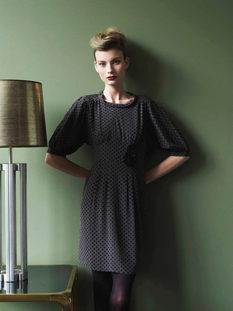 A woman in a vintage-inspired dress poses indoors with soft lighting next to a modern lamp, against a muted green backdrop.