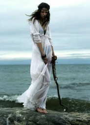 Woman in a long white sheer dress standing on rocks by the ocean holding a wooden staff under an overcast sky.