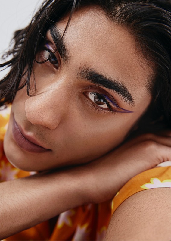 Close-up portrait of a young adult male with dark hair and brown eyes, wearing purple eyeliner and a patterned shirt, resting on his arm.