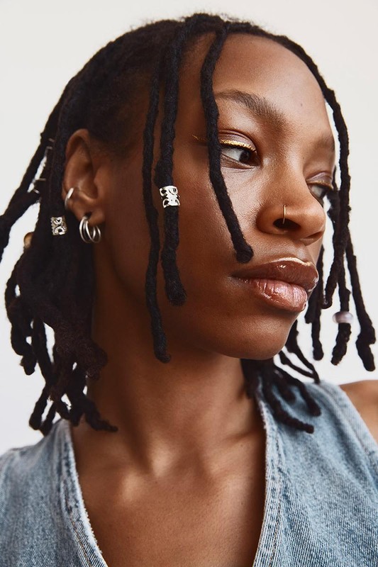 Portrait of a young Black woman with locs decorated with silver beads, wearing a blue denim top against a plain white backdrop.