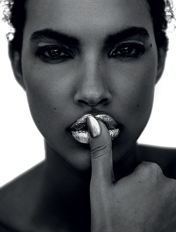 Monochrome close-up of a woman with silver lipstick holding a finger to her lips, set against a bright white background.
