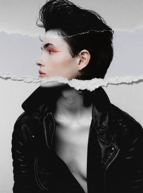 A model with dark hair and orange eyeliner poses in a black leather jacket, framed by ripped paper against a neutral background in studio lighting.