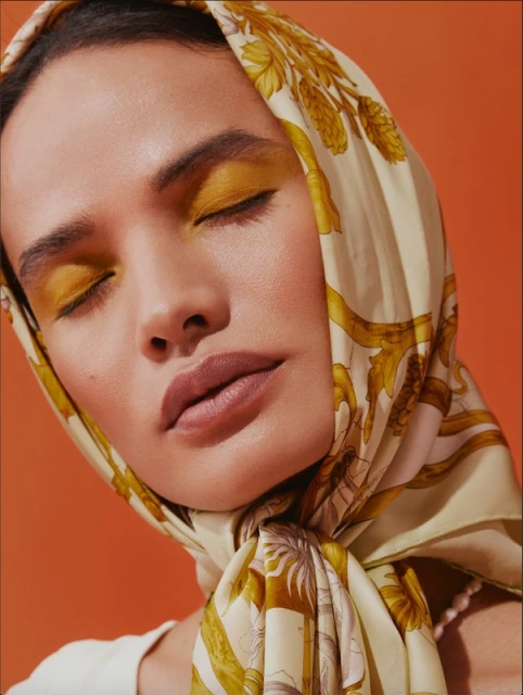 Portrait of a woman with golden eye makeup, closed eyes, and a patterned scarf on an orange background in a beauty fashion shot.