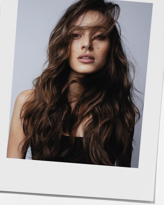 Portrait of a young adult woman with long, wavy brown hair and a black top, styled in a Polaroid-like frame against a neutral background.