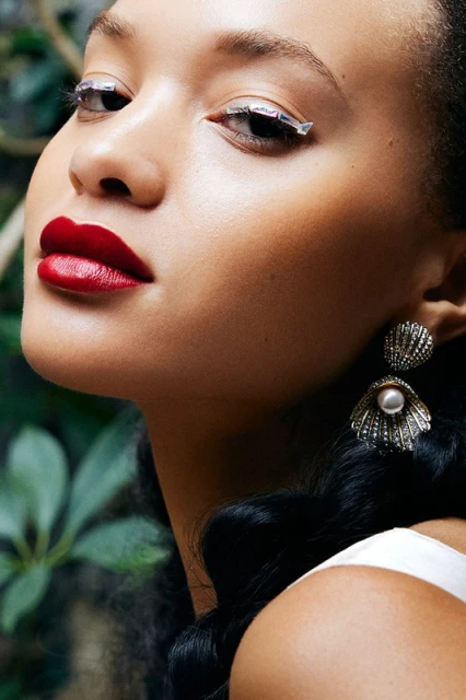 Close-up of a woman with creative silver eye makeup, bold red lipstick, and shell-shaped pearl earrings, set against a leafy green background.
