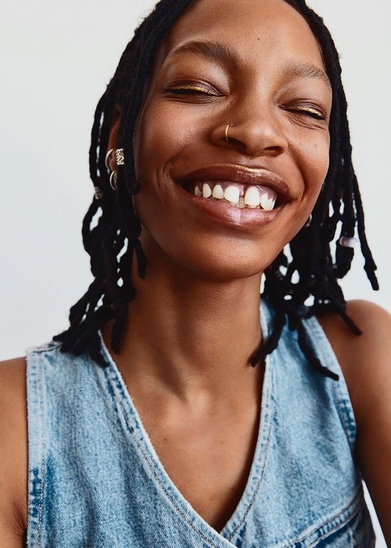Close-up studio shot of a smiling Black woman with dark locs wearing a light-blue denim top and a gold eye shadow against a white background.