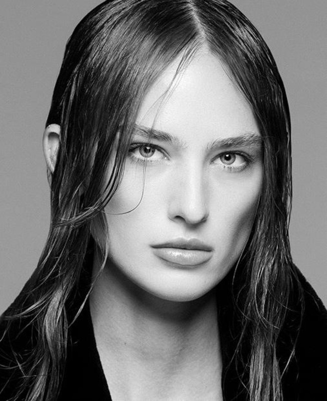 Striking black and white portrait of a woman with wet-look hair, dark clothing, and neutral expression against a soft grey backdrop.