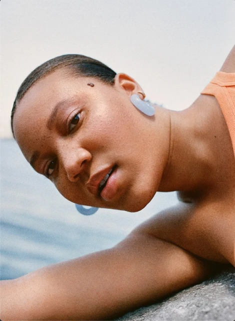 Close-up portrait of an adult woman with shaved head and blue earrings, resting her head on a rock with a light blue sky backdrop.