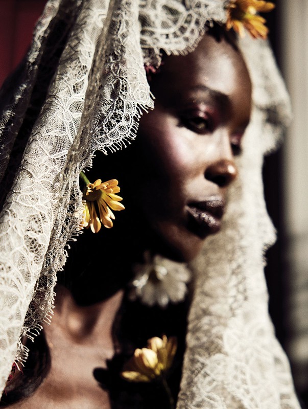 Profile portrait of a dark-skinned woman wearing a lace veil and yellow flowers in her hair and on her chest against a dark red background.