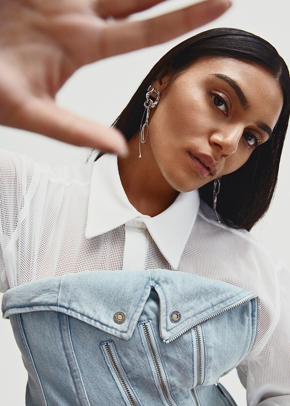 Portrait of a woman in a denim bustier, white top, and silver earrings against a white backdrop, emphasizing fashion and studio lighting.
