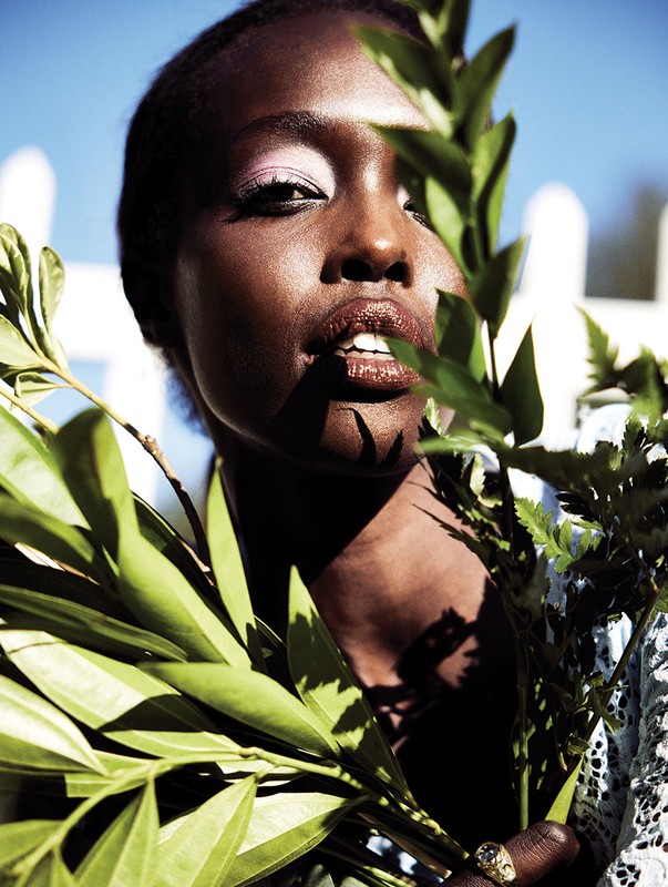Close-up portrait of a black woman with bright pink eye shadow, holding green leafy branches in front of a white picket fence.