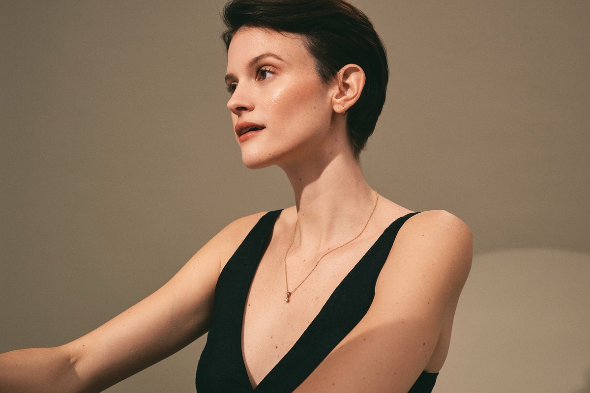 Elegant woman in a black top and gold necklace poses indoors against a grey wall, creating a minimalist beauty and fashion portrait.
