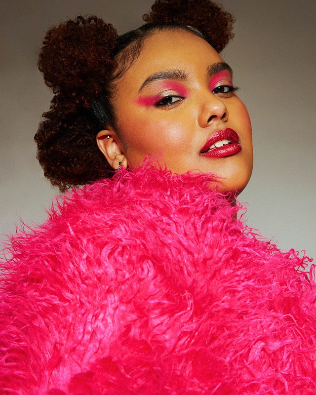 Close-up portrait of a woman with dark curly hair styled in space buns, wearing bright pink makeup and a fluffy pink jacket.