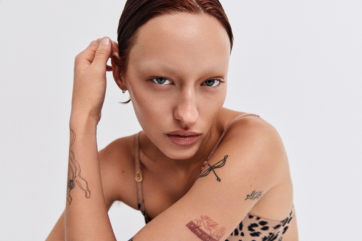 Close-up portrait of a young adult female model with wet red hair and face tattoos against a plain white background in photo studio.