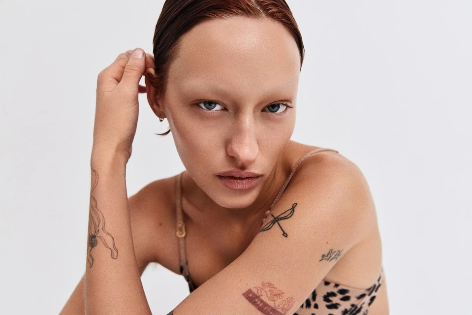 Close-up portrait of a young adult female model with wet red hair and face tattoos against a plain white background in photo studio.