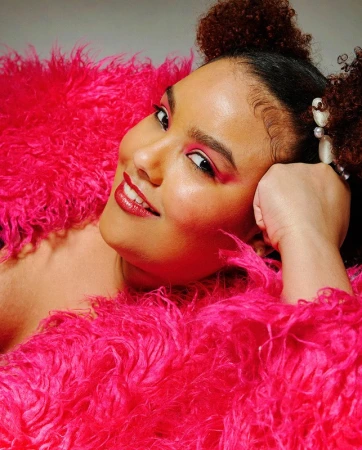 Portrait of a smiling woman with pink eye makeup and curly hair styled in space buns, posing in a bright pink fluffy shawl.