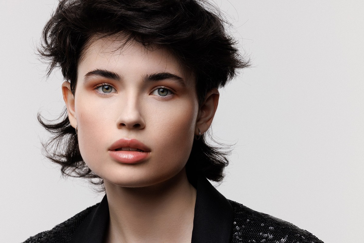 Close-up portrait of a young adult woman with dark, curly hair wearing a black, sparkling sequined jacket on a neutral background.
