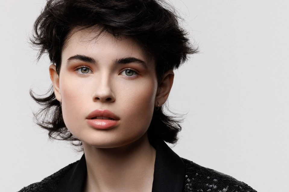 Close-up portrait of a young adult woman with dark, curly hair wearing a black, sparkling sequined jacket on a neutral background.
