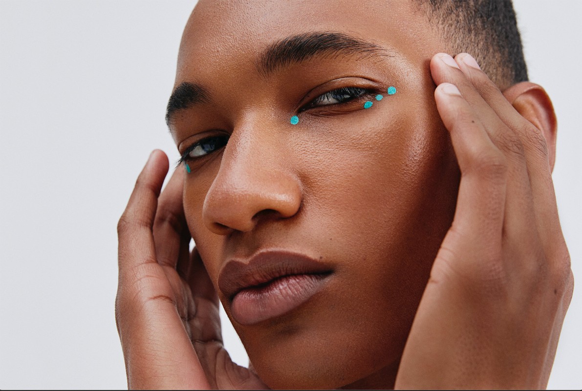 Close-up portrait of a young adult Black man with teal dots around his eyes, hands near his face, set against a plain white backdrop.