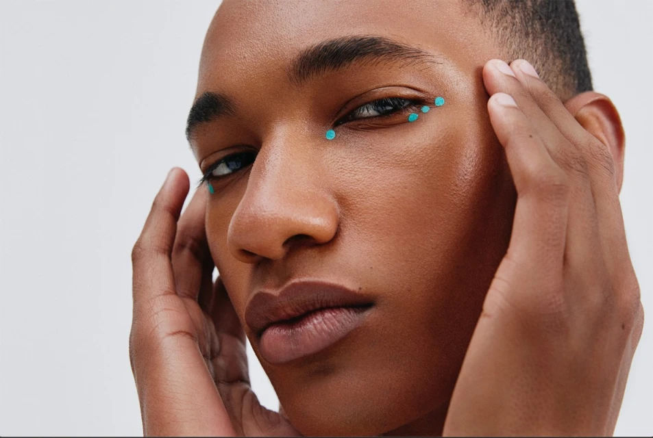 Close-up portrait of a young adult Black man with teal dots around his eyes, hands near his face, set against a plain white backdrop.