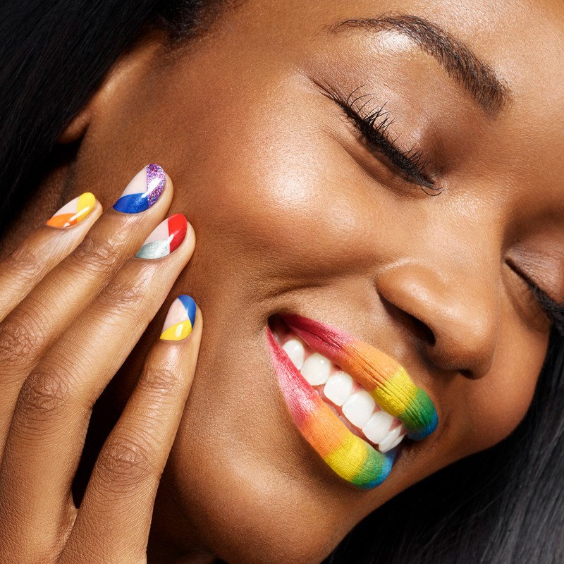 Close-up of a smiling dark-skinned woman with rainbow gradient lipstick and colorful geometric nail art, eyes closed.