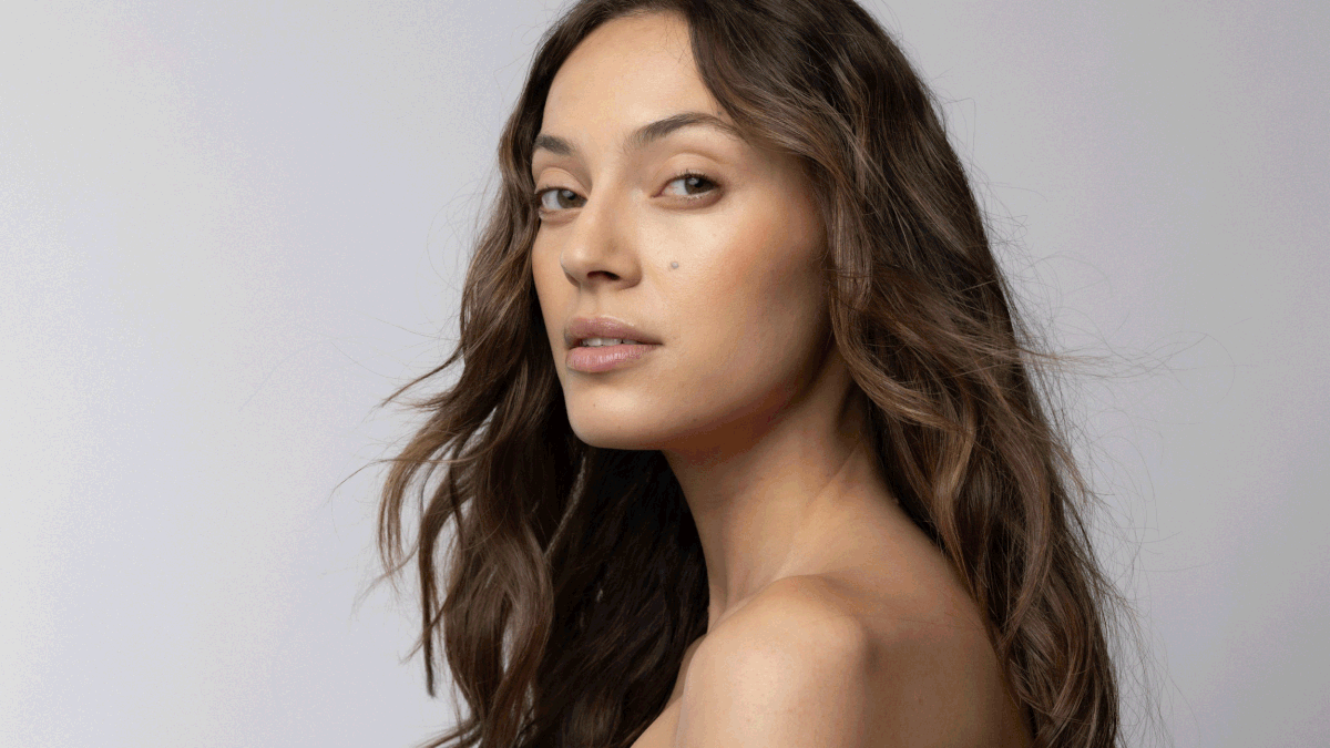 Close-up portrait of a young adult woman with wavy brunette hair in a studio setting, showcasing neutral makeup and natural lighting.