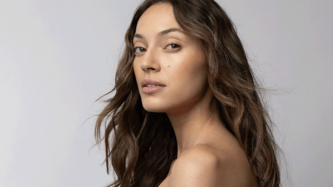 Close-up portrait of a young adult woman with wavy brunette hair in a studio setting, showcasing neutral makeup and natural lighting.