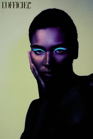 Close-up beauty shot of a person with short black hair and neon blue eyeshadow and violet skin tones, set against a pale green backdrop.