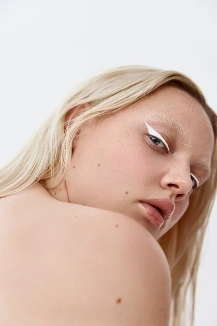 Close-up portrait of a blonde woman with white eyeliner and pale eyebrows against a neutral background, focusing on beauty and makeup.