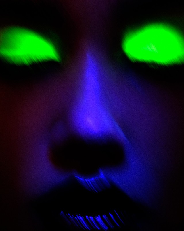 Close-up of a woman's face with glowing green eyes and blue skin, creating a surreal and otherworldly appearance, with dark shadows.