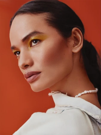 Close up portrait of a woman with yellow eye shadow wearing a white denim jacket and a pearl necklace against a burnt orange backdrop.