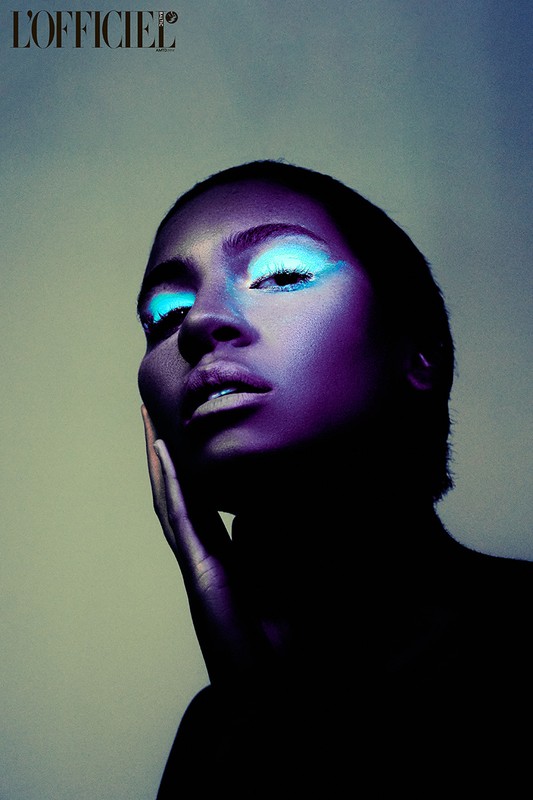 Close-up portrait of a Black adult female model with blue eyeshadow, cool lighting, and hand on face. The magazine L'Officiel logo is visible.