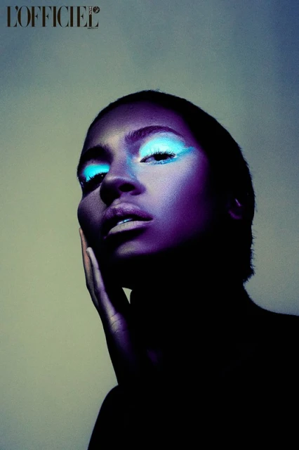 Close-up portrait of a Black adult female model with blue eyeshadow, cool lighting, and hand on face. The magazine L'Officiel logo is visible.