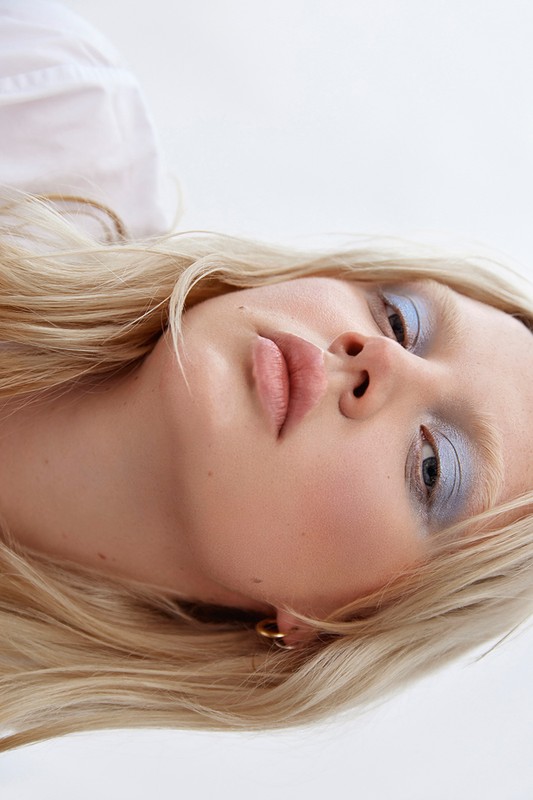 Close-up portrait of a blonde woman wearing blue eyeshadow and gold hoop earrings against a white background.