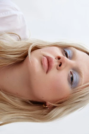 Close-up portrait of a blonde woman wearing blue eyeshadow and gold hoop earrings against a white background.