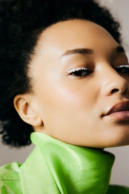 Close-up portrait of a Black woman with pearl makeup, glossy lips, and an apple-green top against a neutral backdrop in soft, diffused lighting.