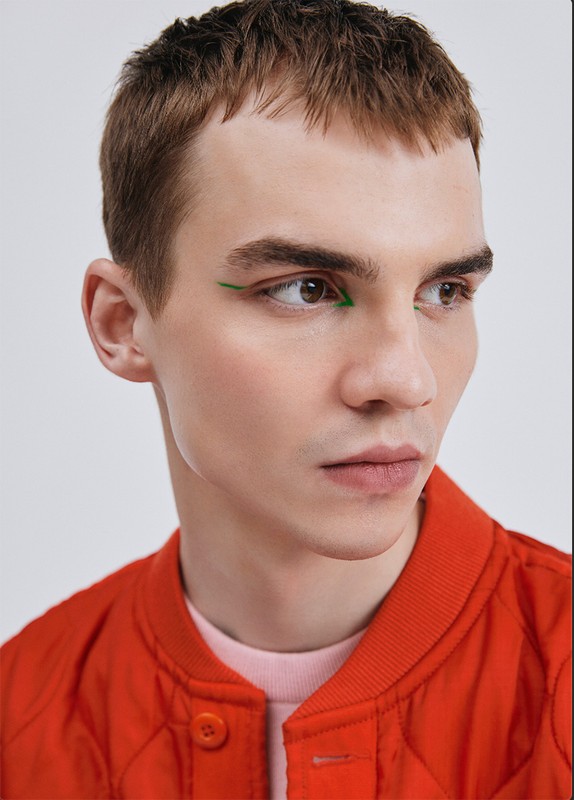Close-up portrait of a young adult male with short brown hair and green eyeliner, wearing a red bomber jacket against a white background.