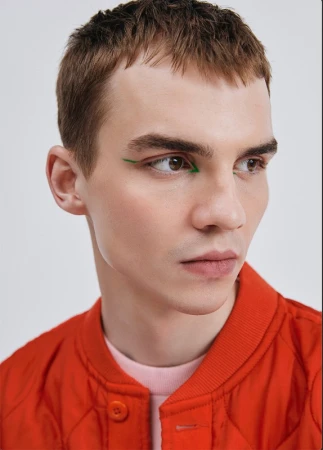 Close-up portrait of a young adult male with short brown hair and green eyeliner, wearing a red bomber jacket against a white background.