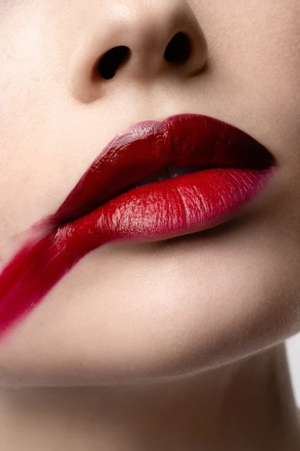 Close-up shot of an adult female face with red lipstick smeared across her upper lip against a pale background, beauty advertisement.