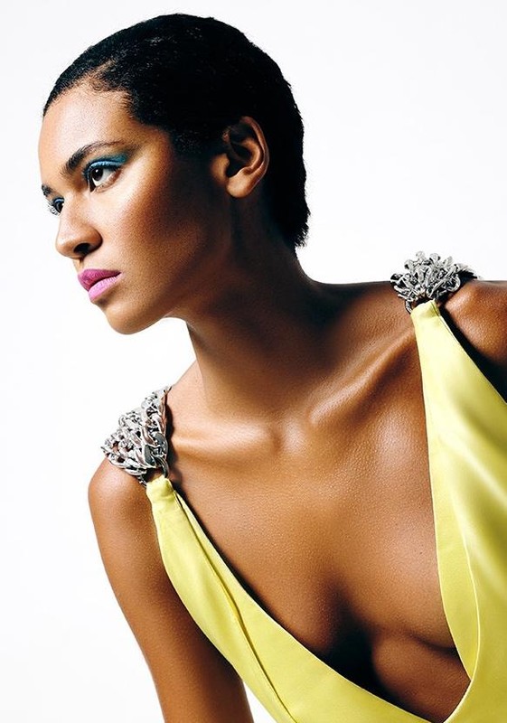 Elegant studio portrait of a Black woman in a yellow dress with jewel-encrusted straps against a bright white backdrop, showcasing fashion and beauty.
