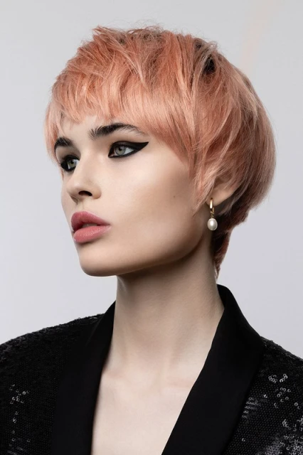 Portrait of a beautiful young woman with short, light pink hair, wearing a black sequin jacket and dangling pearl earrings in a studio shot.